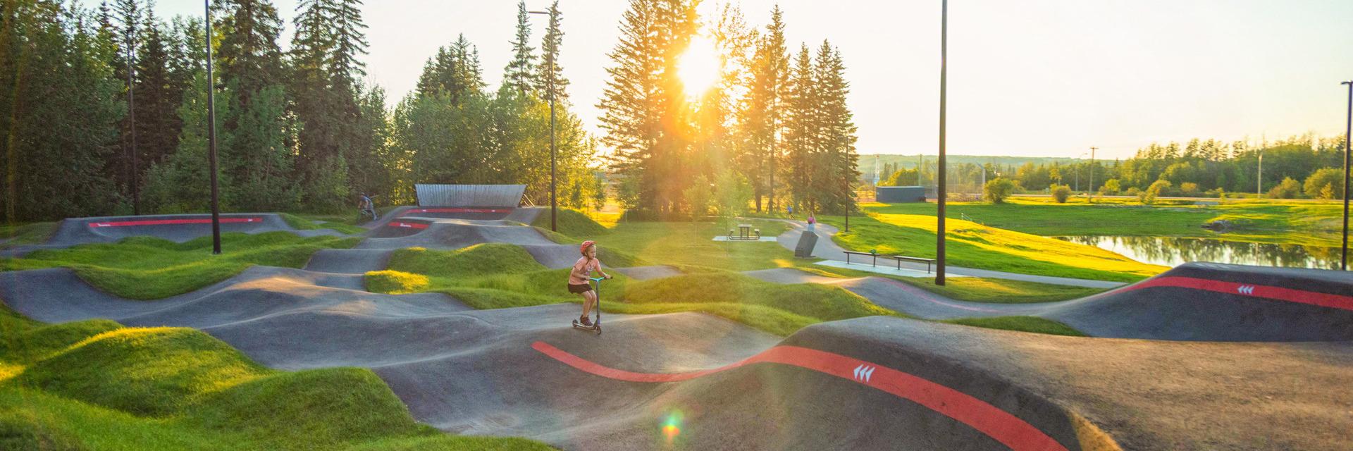 A child rides a scooter around the Kin Park Pump Track on a summer evening.