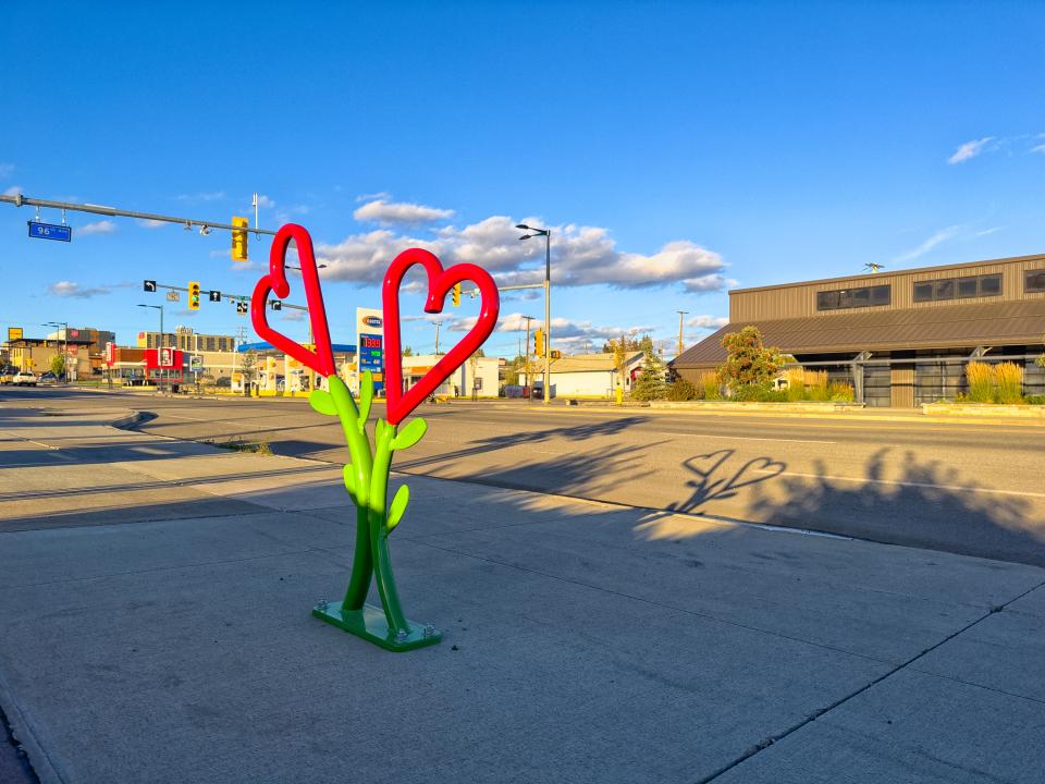 A green bike rack sculpture with red heart-shaped tops casts heart-shaped shadows on the sidewalk.