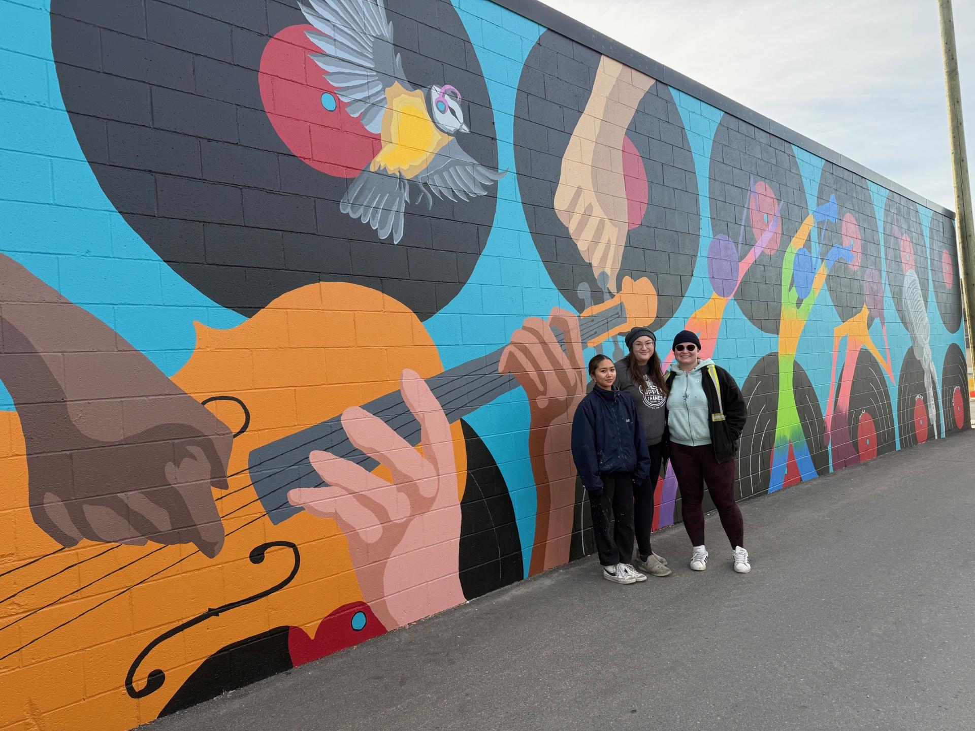 Three artists stand in front of a vibrant mural depicting musical themes, including guitars, records, and a colourful bird.