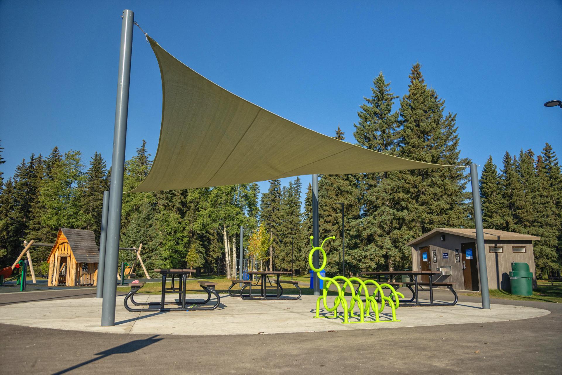 A bright green caterpillar-shaped bike rack sits in Kin Park with trees and a small cabin are in the background.